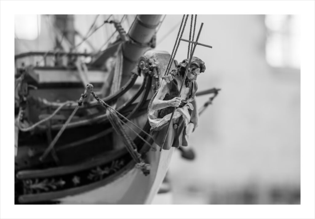 Saint-Jean-du-doigt : Photo en noir et blanc de la figure de proue d'une maquette de bateau détaillée, représentant un saint sculpté tenant une croix à la proue du navire. L'arrière-plan est légèrement flou, ce qui met en valeur l'artisanat et les éléments nautiques au premier plan.
