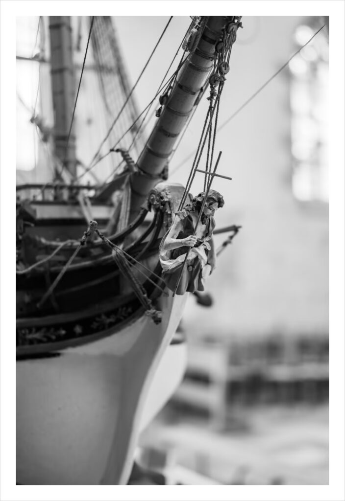 Saint-Jean-du-doigt : Photo en noir et blanc de la figure de proue d'une maquette de bateau détaillée, représentant un saint sculpté tenant une croix à la proue du navire. L'arrière-plan est légèrement flou, ce qui met en valeur l'artisanat et les éléments nautiques au premier plan.