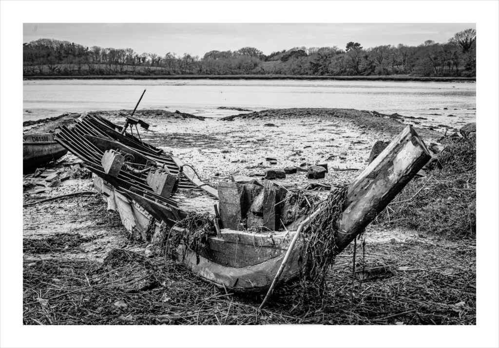 Rivière du Bono : Photo en noir et blanc d'un bateau en bois en décomposition sur un rivage boueux. Le bateau est cassé et envahi par les mauvaises herbes, avec des planches manquantes et des nervures internes visibles. À l'arrière-plan, on aperçoit une eau calme, des arbres lointains et un ciel couvert.