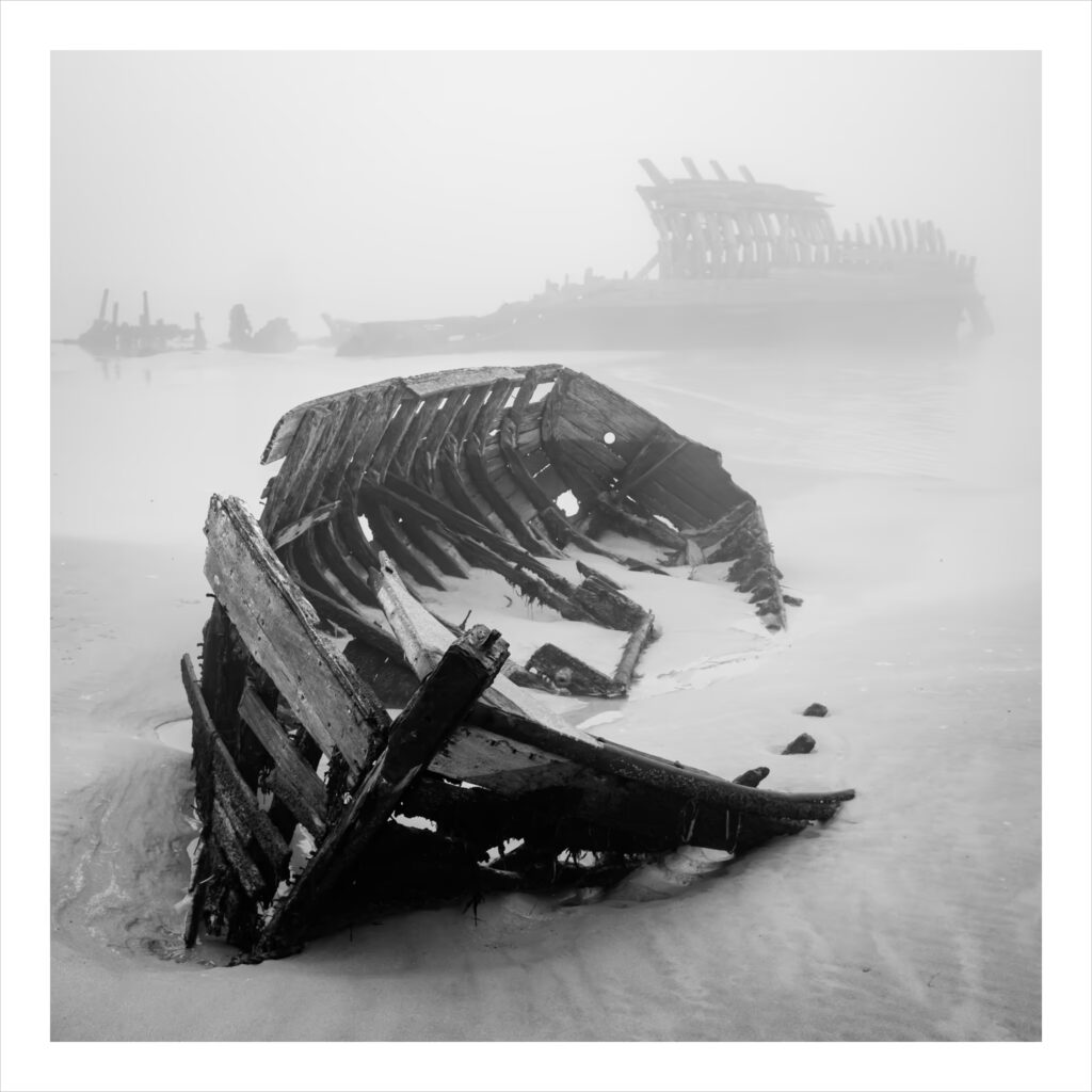 Le Magouer : Deux épaves en décomposition gisent sur une plage brumeuse. Les côtes en bois de l'épave la plus proche sont tordues et partiellement enfouies dans le sable humide, tandis que l'épave la plus éloignée et la plus grande se tient droite dans le lointain brumeux. L'atmosphère est inquiétante et tranquille.