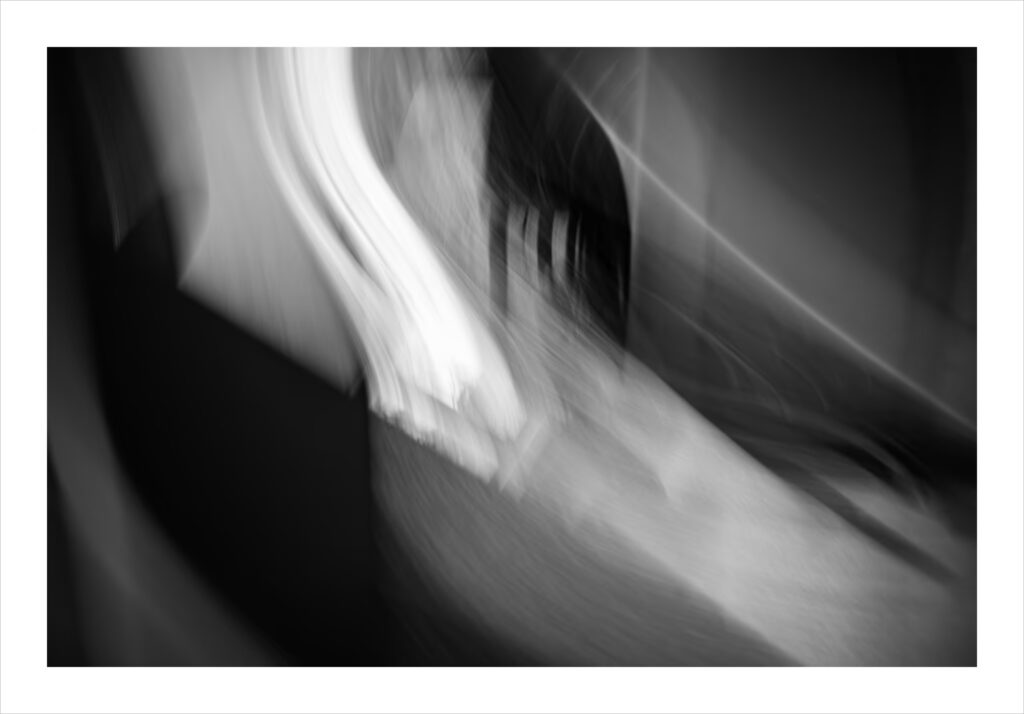 Photo abstraite en noir et blanc avec des lignes floues et des traînées de lumière. Une forme blanche verticale et lumineuse domine le centre gauche, tandis que des formes courbes plus sombres l'entourent. L'effet global est un mouvement dynamique et des formes douces et indistinctes.